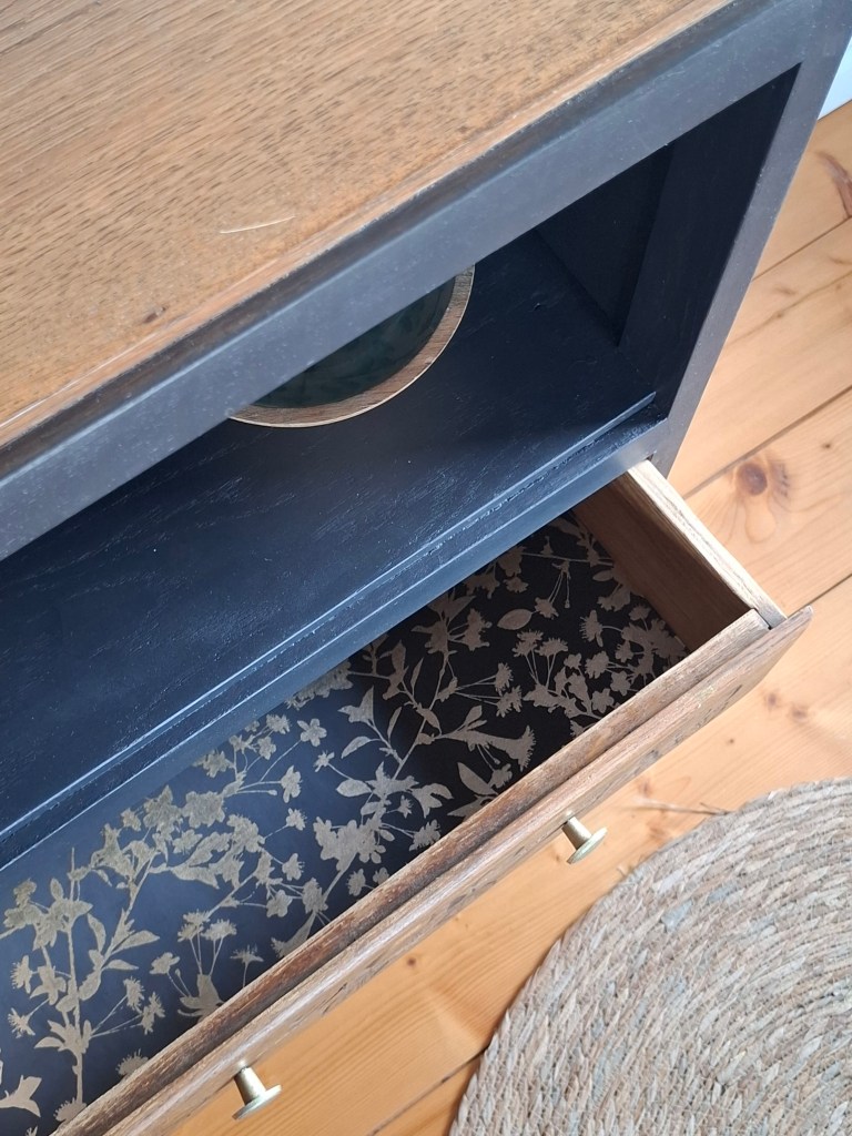 Vue du dessus d'un meuble avec un tiroir ouvert, présentant un fond décoré de motifs floraux dorés sur un fond noir.