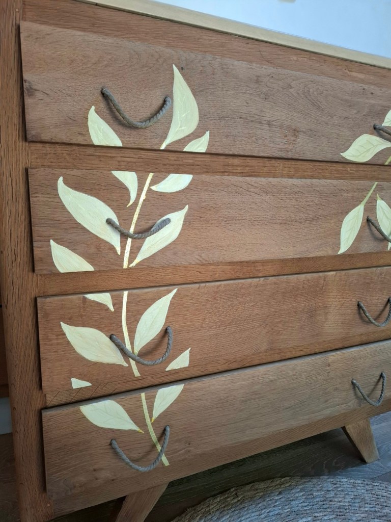 Tiroir en bois avec des motifs de feuilles peintes en crème et des poignées en corde.