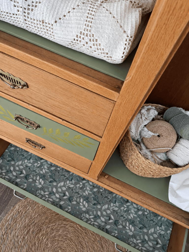 Vue d'un meuble en bois avec des tiroirs, une couverture blanche en crochet sur le dessus, et un tiroir décoré de motifs floraux.