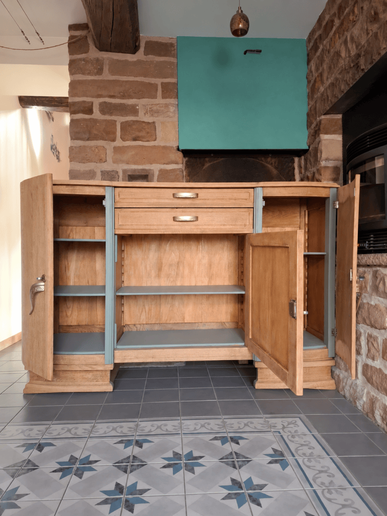 Un meuble de cuisine en bois clair avec des tiroirs et des portes ouvertes, montrant des étagères intérieures. En arrière-plan, un mur en pierre et un plafond bleu.