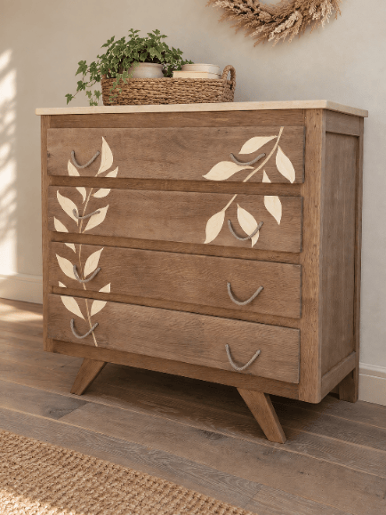 Commode en bois clair avec des décorations de feuilles sur les tiroirs, surmontée d'un plateau en bois et d'un panier de plantes.