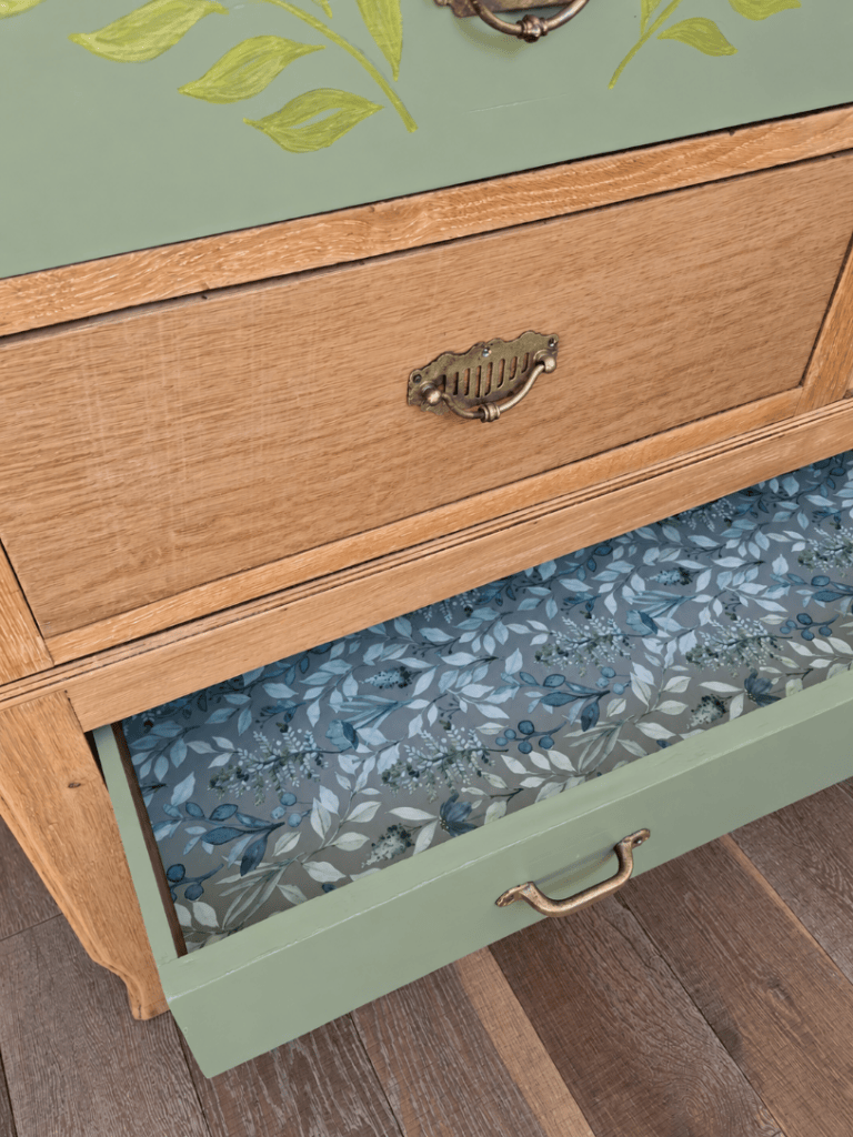Commode en bois avec des tiroirs en bois clair et une façade verte décorée de feuilles, un tiroir est ouvert montrant un intérieur à motif floral bleu.