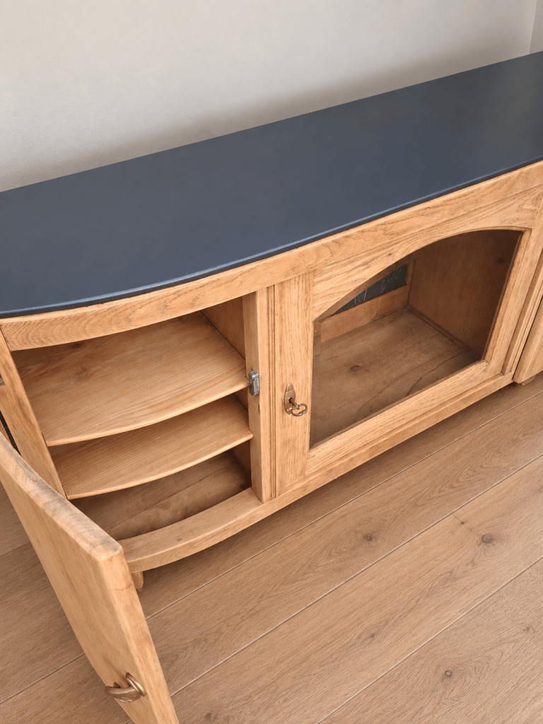 Meuble en bois clair avec un dessus noir, contenant des étagères et une porte vitrée ouverte.
