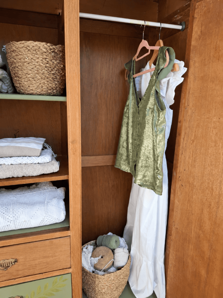 Intérieur d'une armoire en bois avec une robe verte suspendue et une robe blanche. Des paniers en osier contenant des étoffes et des pelotes de laine sont visibles sur les étagères.