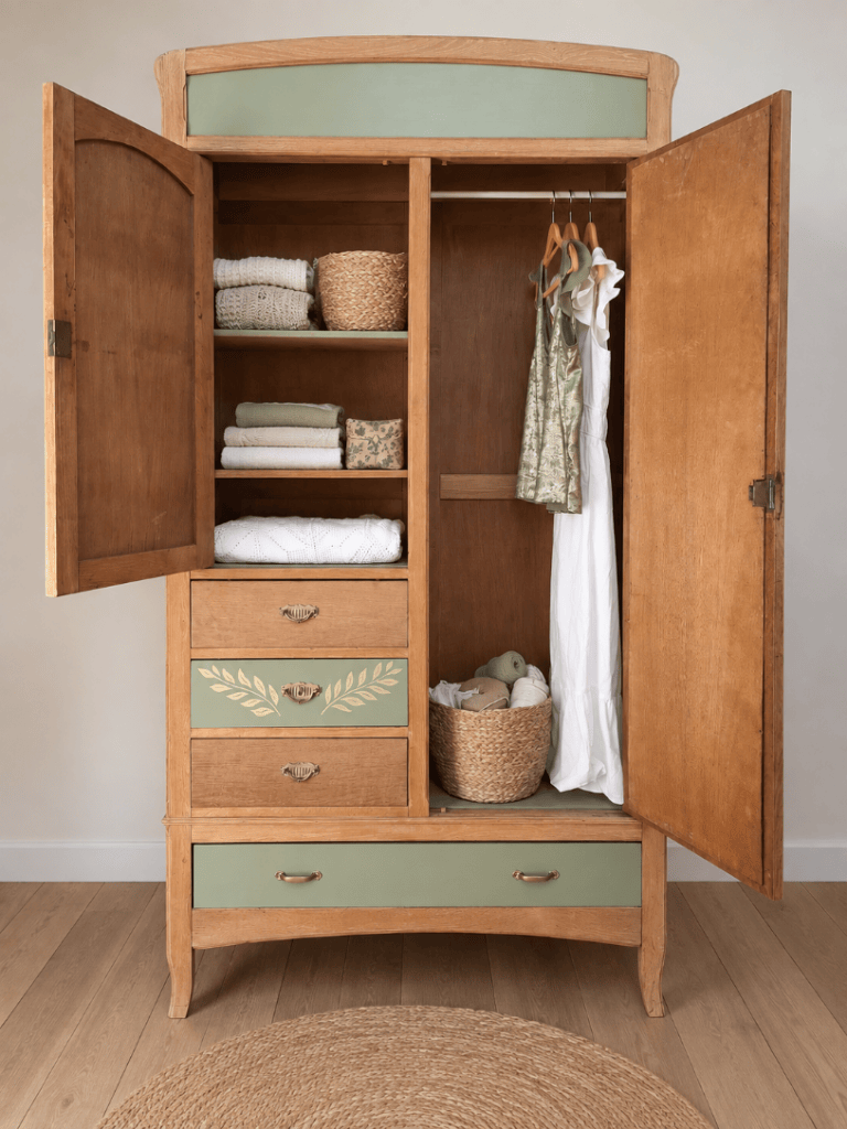 Une armoire en bois ouverte, présentant des étagères avec des serviettes pliées, des paniers en osier et une robe suspendue. Les tiroirs sont également visibles, avec une décoration en feuilles sur l'un d'eux.