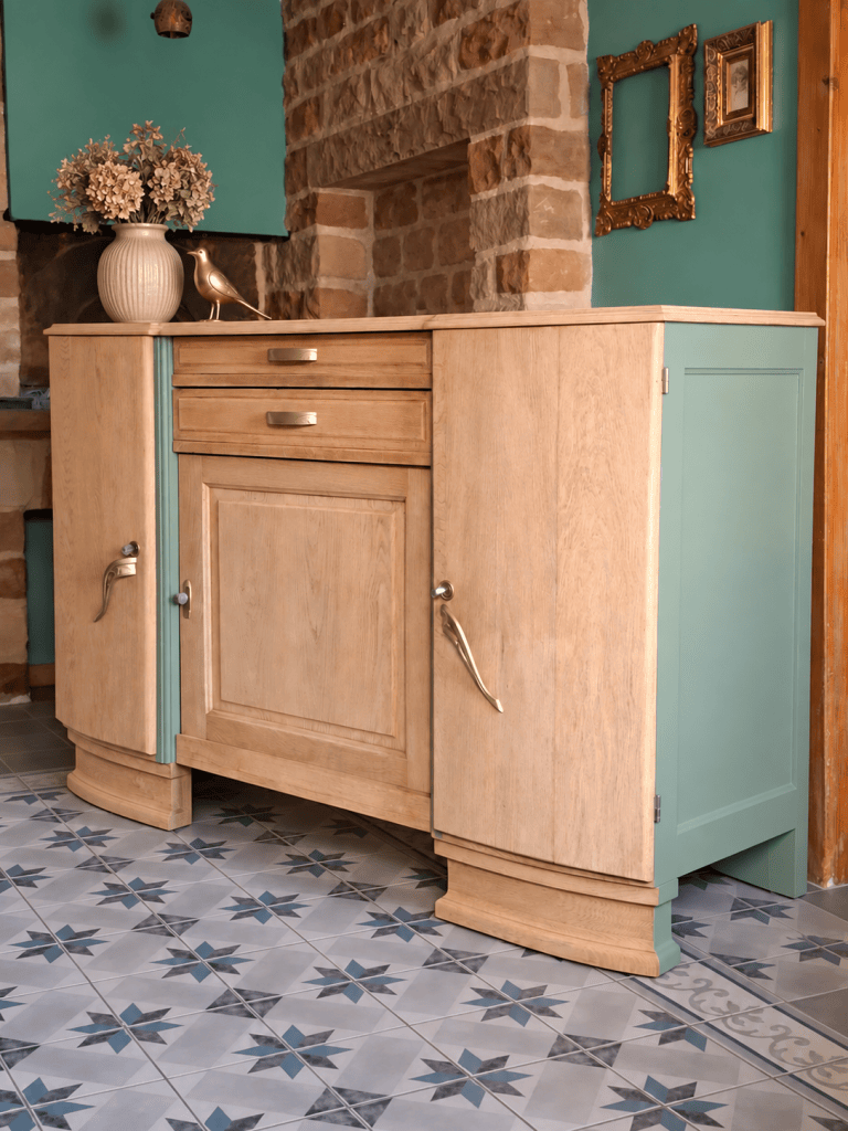 Meuble en bois clair avec des accents turquoise, comprenant une porte centrale, un tiroir et des poignées en métal, situé sur un sol carrelé avec des motifs géométriques.