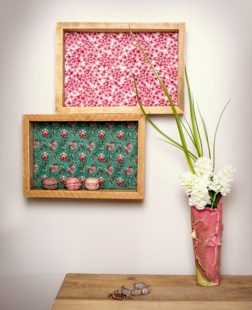 Deux cadres en bois, l'un avec un motif floral rose et l'autre avec un fond vert orné de fleurs, contenant des macarons. Un vase coloré avec des fleurs blanches se trouve à côté sur une table en bois.