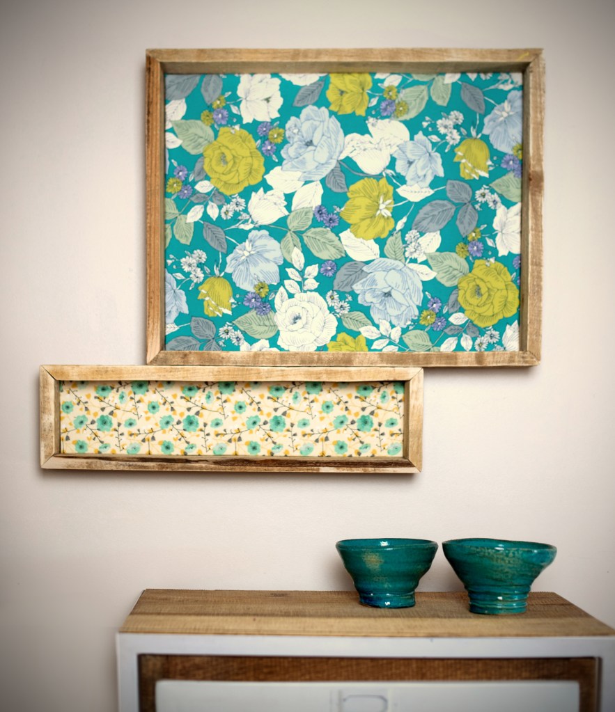 Deux cadres décoratifs avec des motifs floraux accrochés au mur, un cadre supérieur avec des fleurs jaunes et bleues sur fond turquoise, et un cadre inférieur avec des petites fleurs sur fond clair. En dessous, deux bols en céramique verte sont posés sur une table en bois.