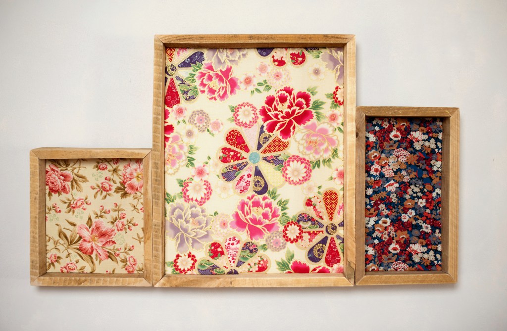 Trois cadres en bois contenant des tissus imprimés floraux aux motifs colorés, accrochés sur un mur blanc.