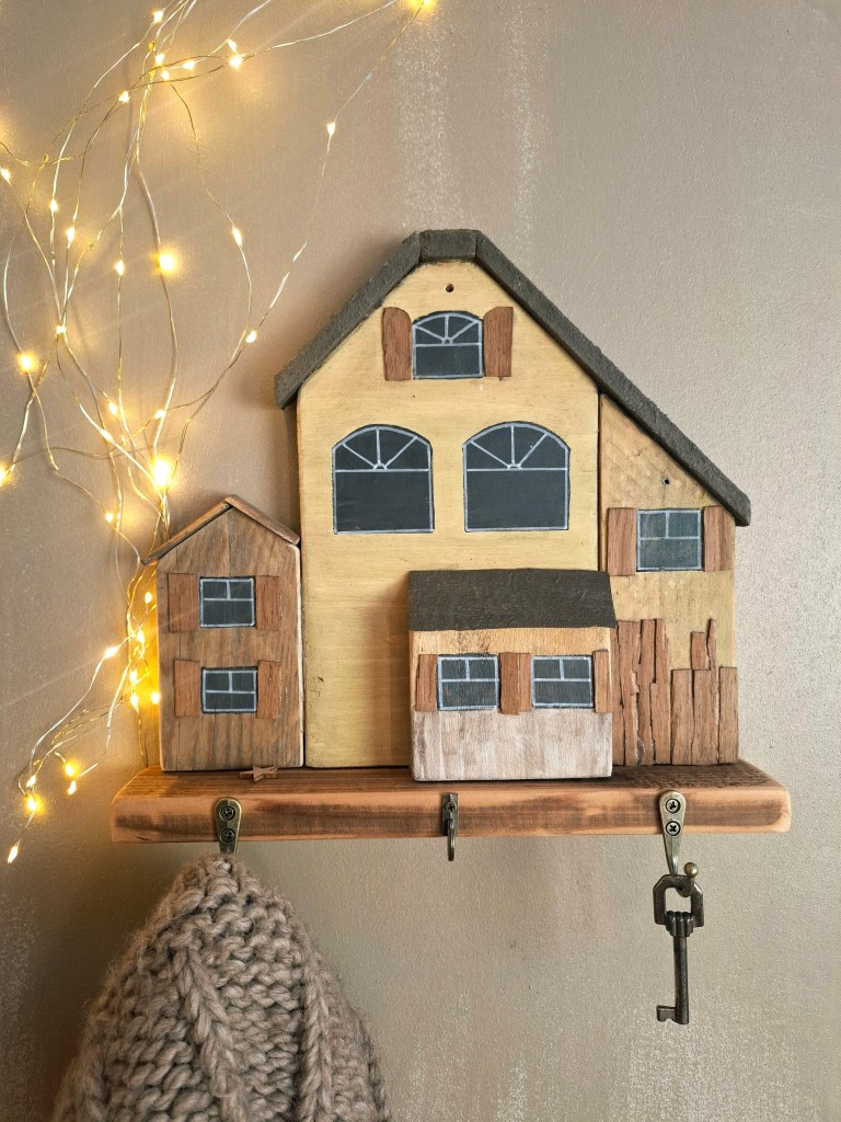 Cadre décoratif en forme de maisons en bois avec trois compartiments, accroché à un mur. Un porte-clé est visible en bas, accompagné de lumières scintillantes.