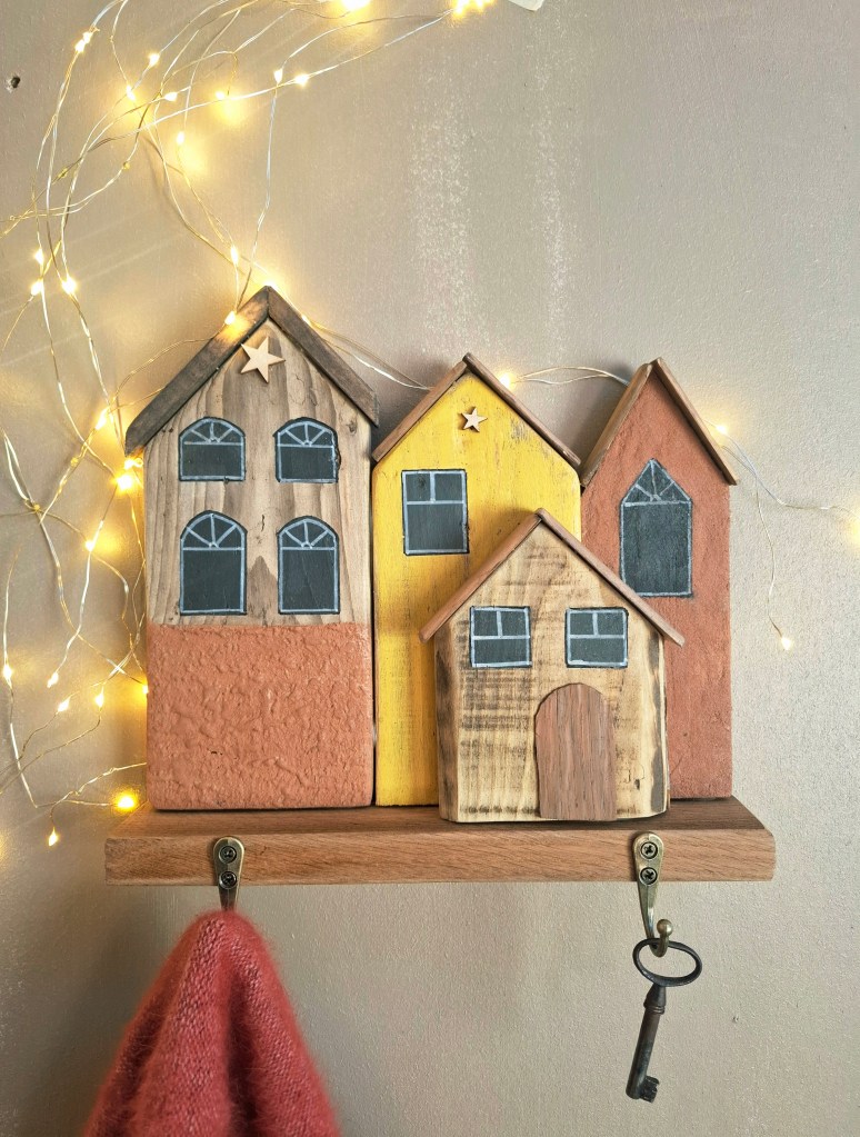 Une décoration murale en bois représentant trois maisons colorées avec des fenêtres dessinées, accrochées à un support en bois avec une clé.