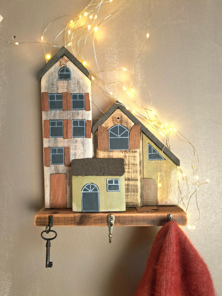 Décor mural en bois représentant trois maisons colorées avec des fenêtres, suspendu sur un mur, accompagné de guirlandes lumineuses. Un crochet en bois en bas pour accrocher des objets.