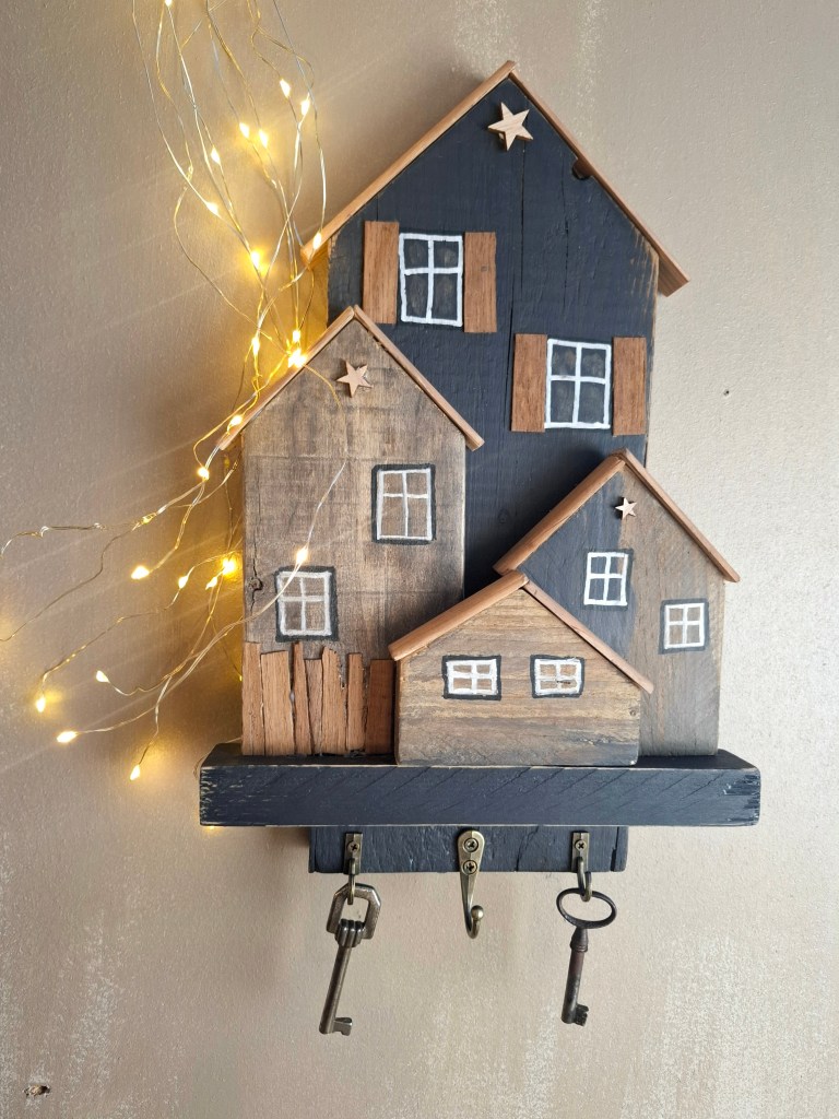 Décor mural en bois représentant des maisons stylisées avec de petites fenêtres et étoiles, accompagné de lumières LED. Une étagère en dessous avec trois crochets pour accrocher des clés.