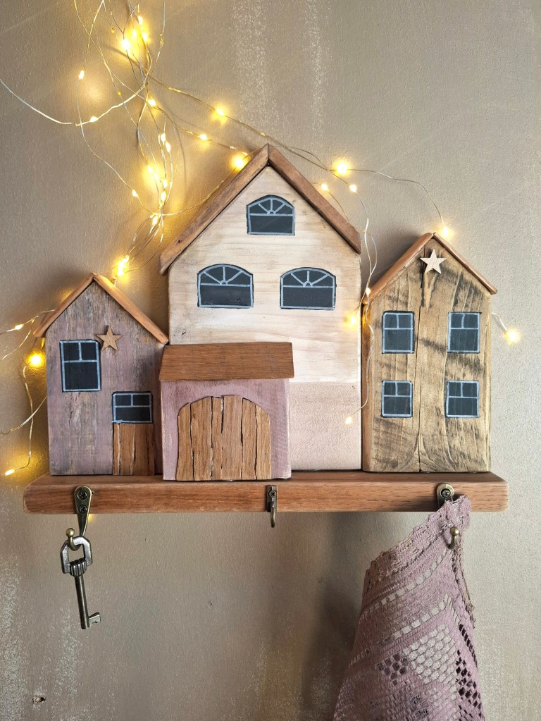 Décoration murale en bois représentant trois maisons stylisées, avec des fenêtres peintes, accrochées sur un support en bois. Une clé est suspendue à un crochet en dessous, entourée de guirlandes lumineuses.