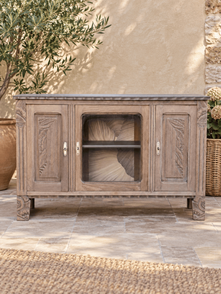 Buffet en bois sculpté avec des portes à vitres, placé sur un sol en pierres, entouré de plantes et d'une mousse décorative.