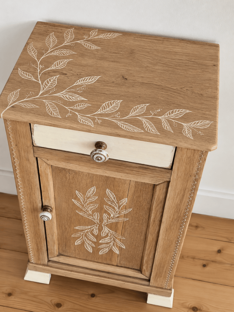 Un meuble en bois avec un dessus orné de motifs de feuilles blanches, comportant un tiroir et une porte, sur un sol en bois clair.