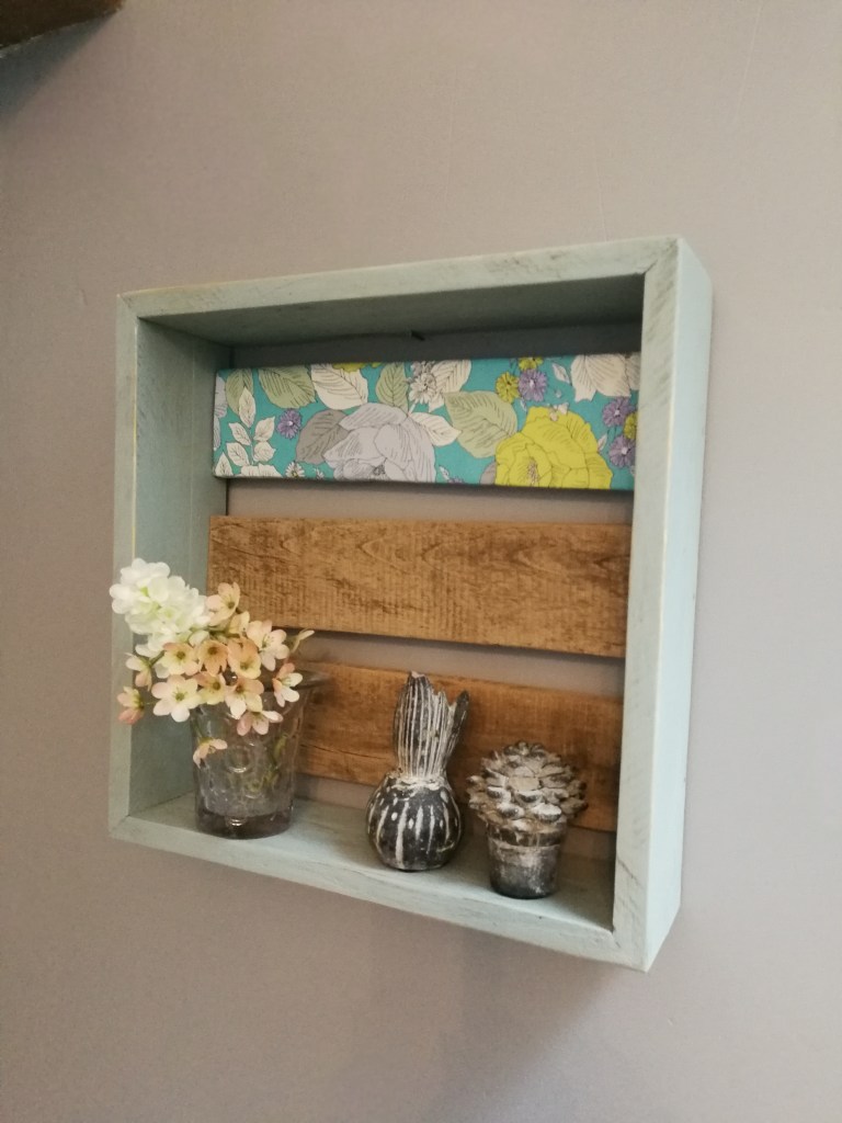 Étagère murale en bois avec décoration incluant des fleurs en pot, des cactus et un fond coloré à motifs floraux.