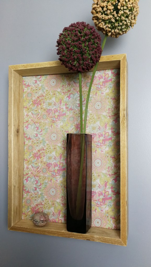 Vase en verre foncé avec des fleurs artificielles dans un cadre en bois, sur un fond de papier peint floral coloré.
